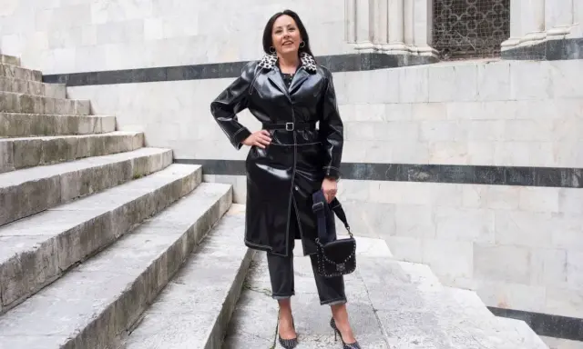 Donna con un audace outfit rock: trench in ecopelle nera, jeans scuri, décolleté borchiate e borsa a tracolla.