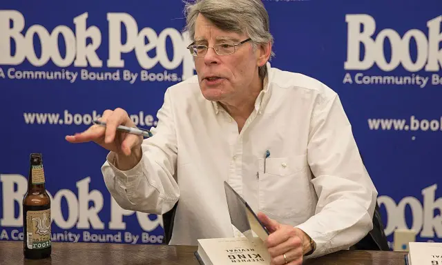 Stephen King, autore di libri da brivido, firma un volume. Sul tavolo, una bottiglia di root beer.