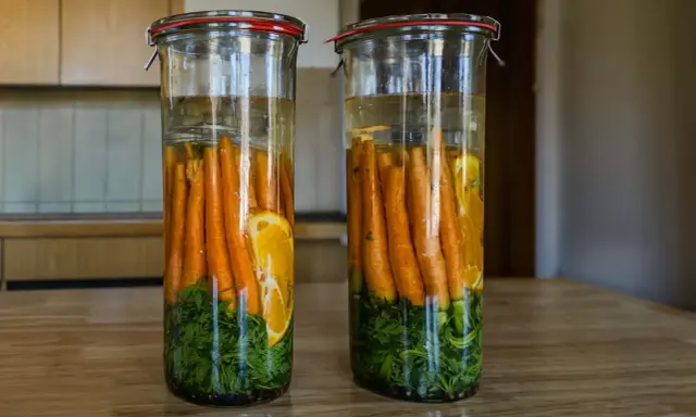 Zwei Gläser mit Karotten, Kräutern und Orangenscheiben in Salzlake zum Fermentieren.
