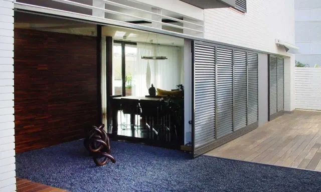 Una moderna casa con una contraventana corredera de lamas metálicas que protege un comedor interior.