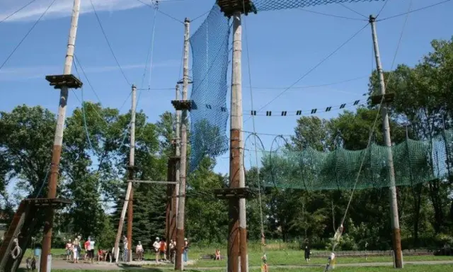 Śląski park linowy z drewnianymi słupami, siatkami i platformami na wysokości. Ludzie przygotowują się do zabawy.