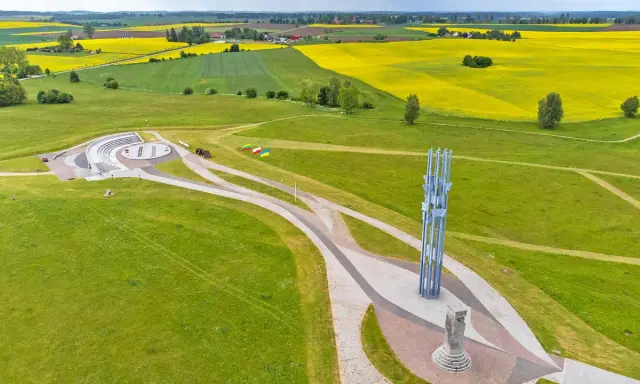 Pomnik i amfiteatr na polach grunwaldzkich, otoczone zielonymi łąkami i kwitnącymi polami rzepaku.