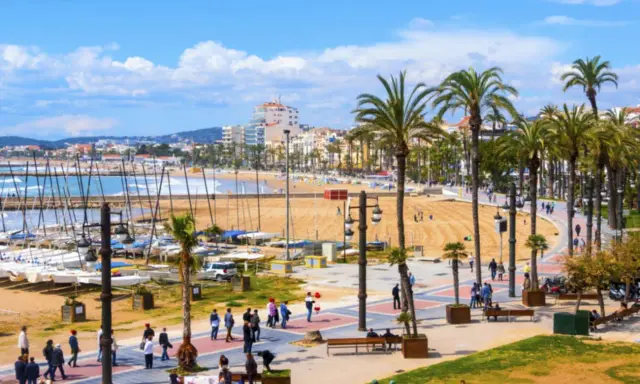 En livlig strandpromenad i Sitges, Spanien, med palmer, en sandstrand, båtar i hamnen och människor som njuter av solen.