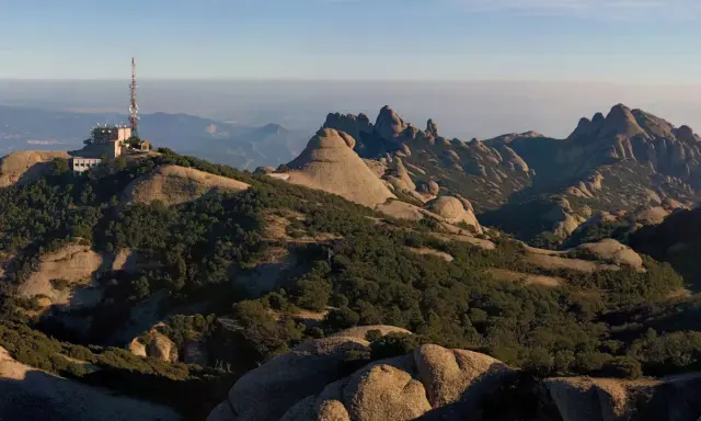 Edificio con antena en la cima de una montaña rocosa, con vistas a un paisaje montañoso y brumoso que se extiende hasta el horizonte, evocando la sensación de estar en planos cerca de Barcelona.