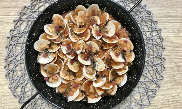 Almejas con jamón en una paellera, con trocitos de jamón y perejil picado, sobre un mantel de madera.