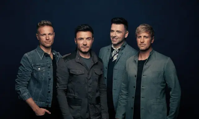 Medlemmar av Westlife poserar för en studiofotografering. De bär moderna jackor och ser direkt in i kameran.
