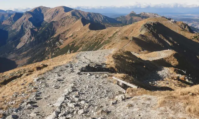 Kamienista ścieżka prowadzi przez trawiaste zbocza, ukazując najpiękniejsze doliny Zakopane. W oddali majaczą góry.