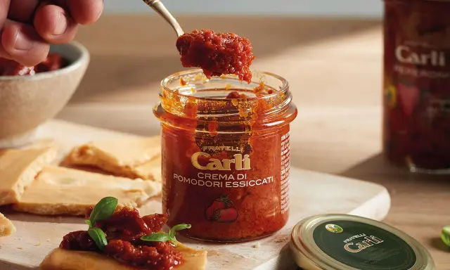Une cuillère prélève de la crème de tomates séchées d'un pot Carli. Servie sur des crackers avec du basilic.