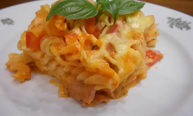 Pyszna zapiekanka z szynką, makaronem fusilli, pomidorami i ciągnącym się serem, ozdobiona listkami bazylii.