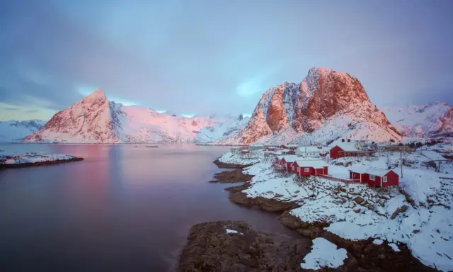 Vackert lofoten vinterlandskap med snöklädda berg, röda hus vid vattnet och en rosa himmel.