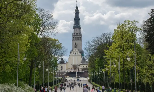 Aleja prowadząca do imponującej wieży klasztornej, jednego z ważnych sanktuariów w Polsce. Ludzie spacerują, podziwiając architekturę i otaczającą zieleń.