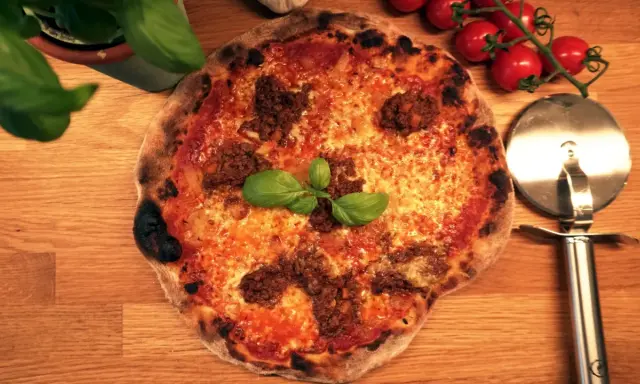 En napolitansk pizza med köttfärs som topping, garnerad med färsk basilika. Bredvid ligger en pizzaskärare och körsbärstomater.