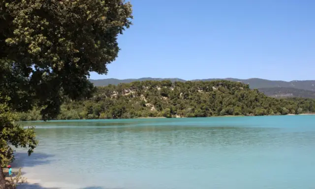 Vue paisible de l'étang de la Bonde, avec ses eaux turquoise bordées de verdure et de collines boisées sous un ciel bleu clair.