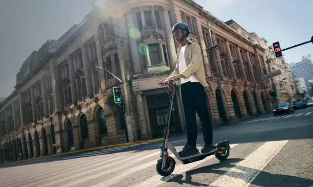 Un homme en casque roule sur la trottinette électrique la plus rapide du monde, traversant une rue bordée de bâtiments historiques.