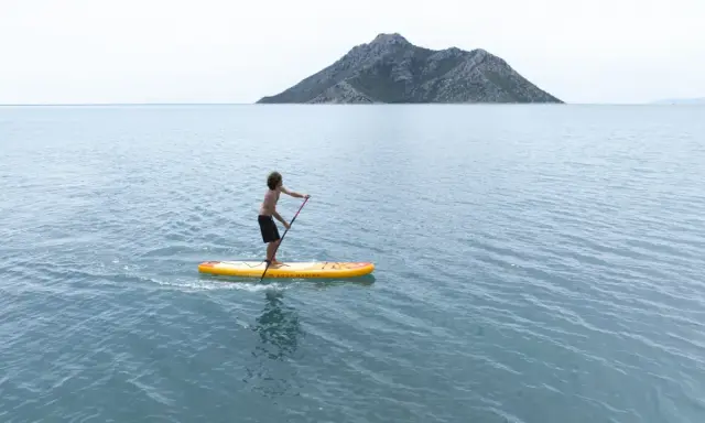 Chłopak na desce sup Aqua Marina przemierza spokojne morze w kierunku skalistej wyspy.