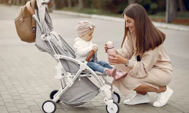 Mama karmi córeczkę z butelki, siedząc obok wózka. Idealne wózki dla dziewczynki na spacer.