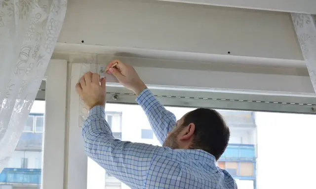 Hombre con camisa a cuadros midiendo para reparar persianas.
