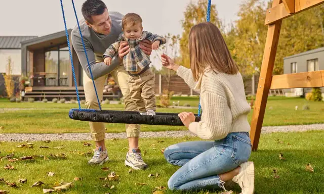 Radosne zabawy w ogrodzie: tata podtrzymuje dziecko na huśtawce, mama zachęca do zabawy.