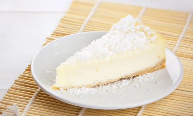 Une part de tarte noix de coco, crémeuse et généreusement saupoudrée de noix de coco râpée, repose sur une assiette blanche, prête à être dégustée.