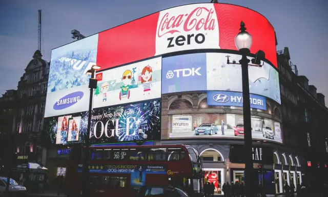 Wielkie ekrany reklamowe na placu, prezentujące różne rodzaje reklamy: Coca-Cola Zero, Samsung, Vogue, Hyundai, TDK. Widać też autobus i ludzi.