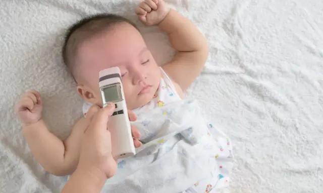 Jak mierzyć temperaturę u niemowlaka termometrem bezdotykowym? Ręka trzyma termometr przy czole śpiącego maluszka.