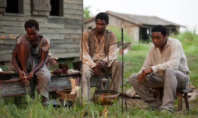 Tre persone sedute all'aperto, in un'ambientazione che ricorda i film biografici sulla schiavitù.