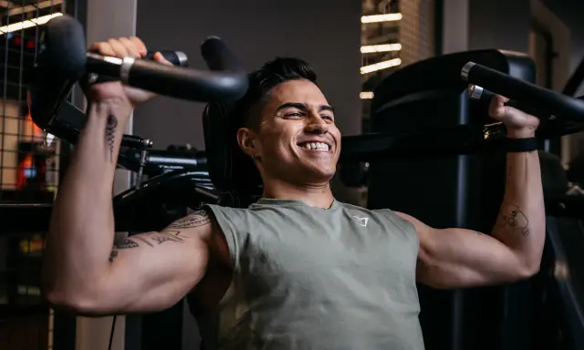 Ein Mann trainiert im Fitnessstudio, um seinen Brustmuskel aufzubauen. Er lächelt zufrieden, während er die Gewichte hebt.