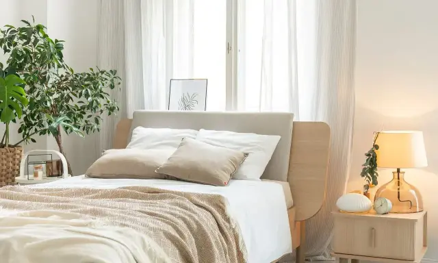Przytulna sypialnia inspiracje: łóżko z beżowym zagłówkiem, biała pościel, narzuta w jodełkę, rośliny, lampka nocna.