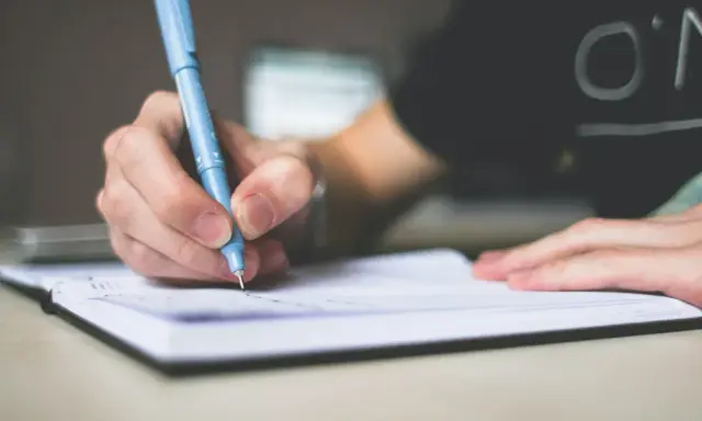 Dłoń pisze niebieskim długopisem w notatniku. To może być początek pisania opinii, esej przykład.