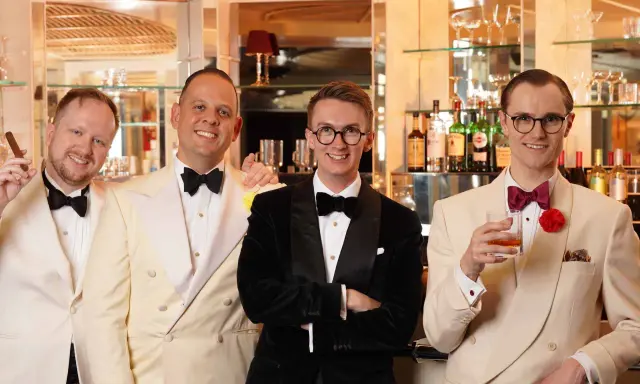 Four men in stylish suits and bow ties, embodying creative black tie, pose at a bar. One holds a cigar, another a drink.