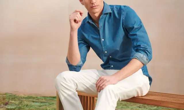 A man poses in a blue denim shirt and crisp white jeans, looking relaxed on a wooden bench.
