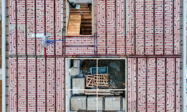 Siatka rur do ogrzewania podłogowego ułożona co 10-15 cm na betonowej płycie fundamentowej. Widoczne schody i materiały budowlane.
