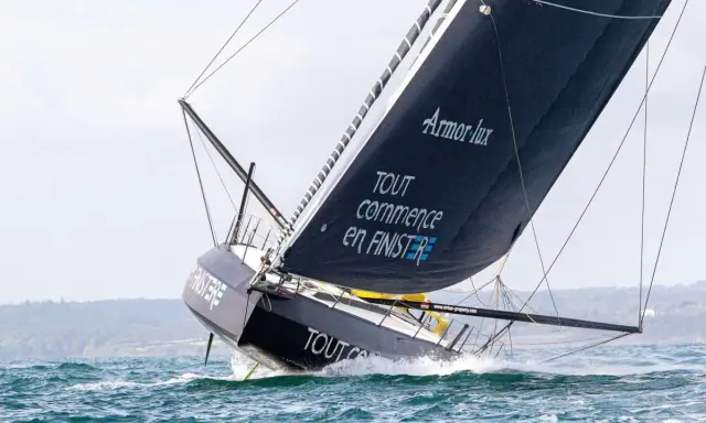 Le voilier IMOCA de Jean Le Cam fend les vagues, sa grand-voile noire ornée du slogan "Tout commence en Finistère" gonflée par le vent.