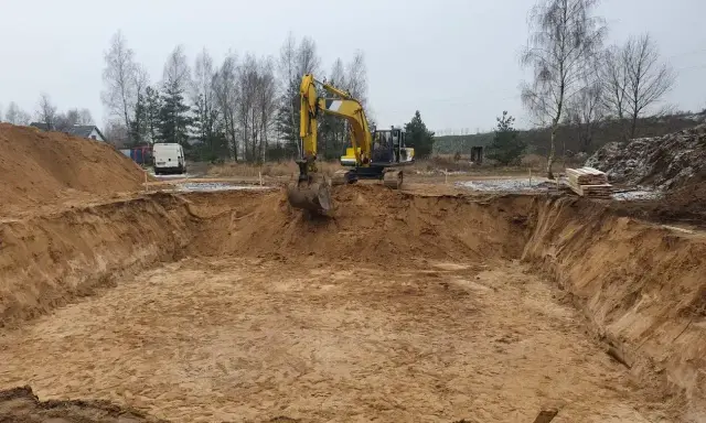Żółty koparka pogłębia wykopy pod fundamenty. W tle drzewa i biały van.