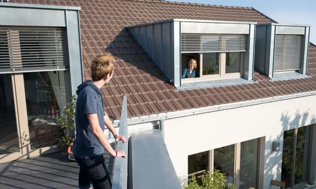 Junge Person auf Balkon blickt zu Frau in Schleppgaube. Verschiedene Dachgauben Arten sind zu sehen.