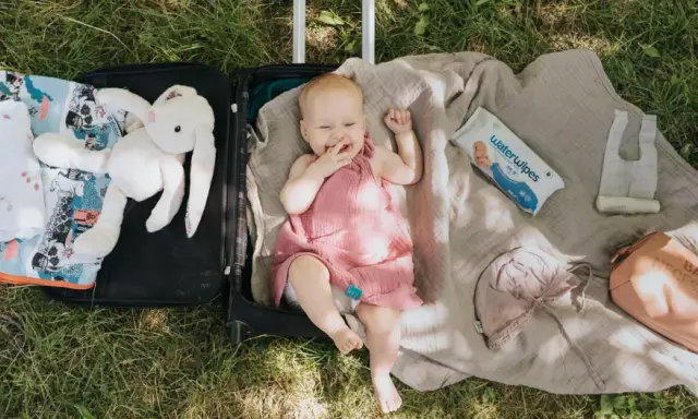 Uśmiechnięte niemowlę w walizce, gotowe na podróże z niemowlakiem. Obok pluszowy króliczek, kocyk, kosmetyczka i chusteczki.