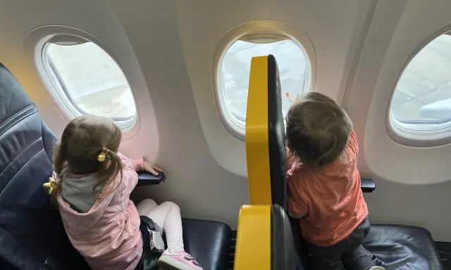Dzieci patrzą przez okno samolotu Wizz Air. Dziewczynka w różowej bluzie i chłopiec w pomarańczowej koszulce, siedzą obok siebie.