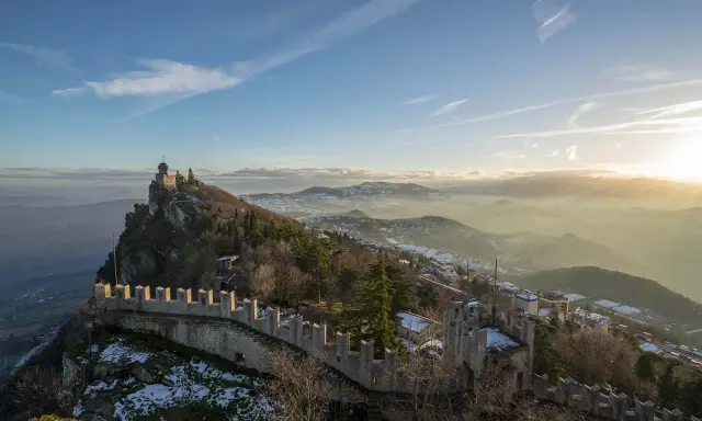 Un suggestivo viaggio a San Marino: torri medievali su un crinale roccioso, con la nebbia che avvolge le valli sottostanti al tramonto.