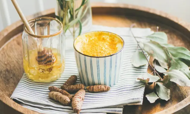 Aromaticzne złote mleko w kubeczku, obok słoiczek z miodem i korzenie kurkumy. Idealne na chłodne wieczory.
