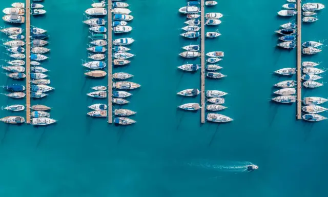 Vue aérienne d'un port où de nombreux voiliers sont amarrés. Un petit bateau navigue, laissant une trace dans l'eau. Le **bloc côtier obligatoire** est bien visible.