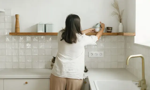 Kobieta układa kubki na drewnianych, otwartych półkach w kuchni. Obok kotek obserwuje porządki.