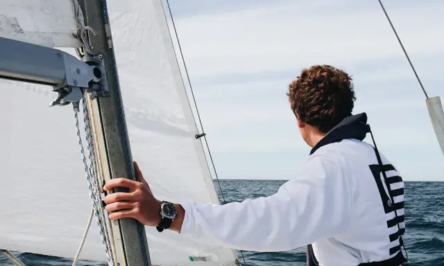 Un jeune homme regarde l'horizon depuis un voilier autonome. La voile blanche est gonflée par le vent.