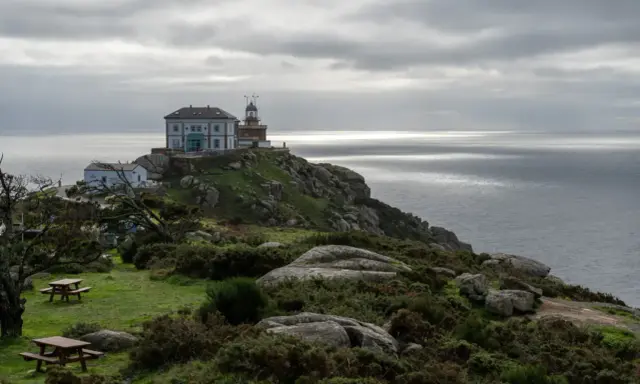 Leuchtturm und Gebäude auf felsiger Küste bei Finisterre, Spanien. Grauer Himmel über dem Meer.
