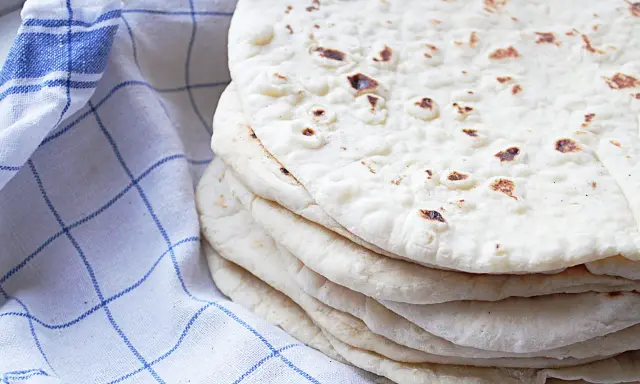 En hög med nybakade tortillabröd, perfekta för ditt nästa tortilla recept. Bredvid ligger en blåvitrutig kökshandduk.