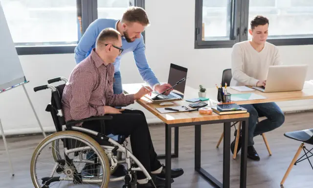 Współpraca w biurze: mężczyzna na wózku inwalidzkim, z umiarkowanym stopniem niepełnosprawności, pracuje z kolegą przy laptopie.