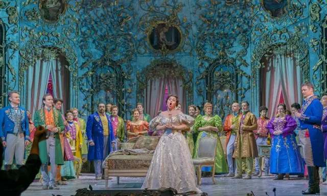 Spiewak operowy w białej sukni śpiewa na scenie, otoczony przez barwnie ubranych artystów w bogato zdobionej scenografii.