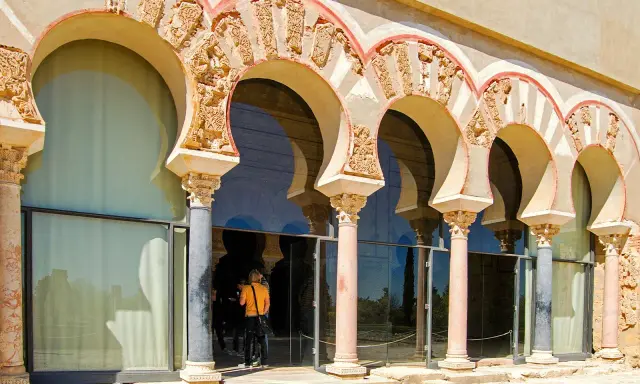 Verzierte Bögen und Säulen der Medina Azahara spiegeln sich in Glas. Besucher bestaunen die historische Architektur.
