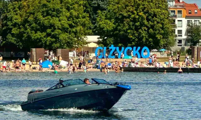 Motorówka sunie po wodzie, w tle plaża pełna ludzi i wielki napis "GIŻYCKO". Idealny dzień na Mazurach.