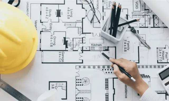 Un arquitecto o proyectista de una obra revisa planos detallados de una casa, con casco amarillo, lápices y calculadora.