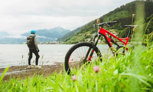 En person med hjälm och ryggsäck står vid en sjö med en mountainbike framför sig. Berg och en stad syns i bakgrunden. Perfekt för att cykla MTB.